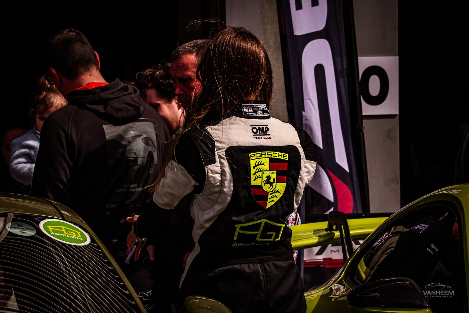 A racing driver wearing a Porsche-branded suit stands near a lime green race car at a motorsport event in dim lighting.