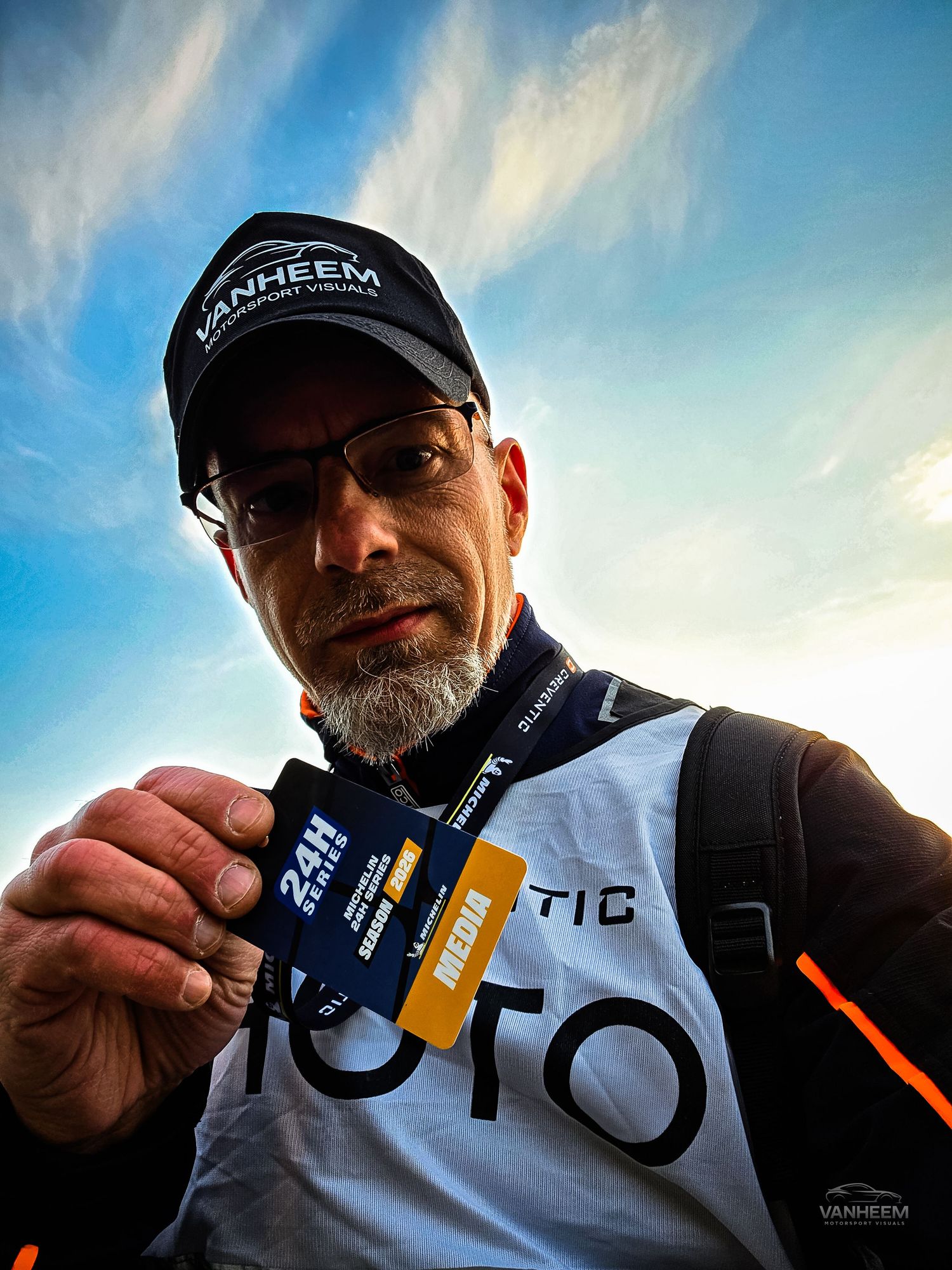 Man holding a 24H media pass at a motorsport event, wearing a Vanheem cap and TOTO vest under a blue sky.