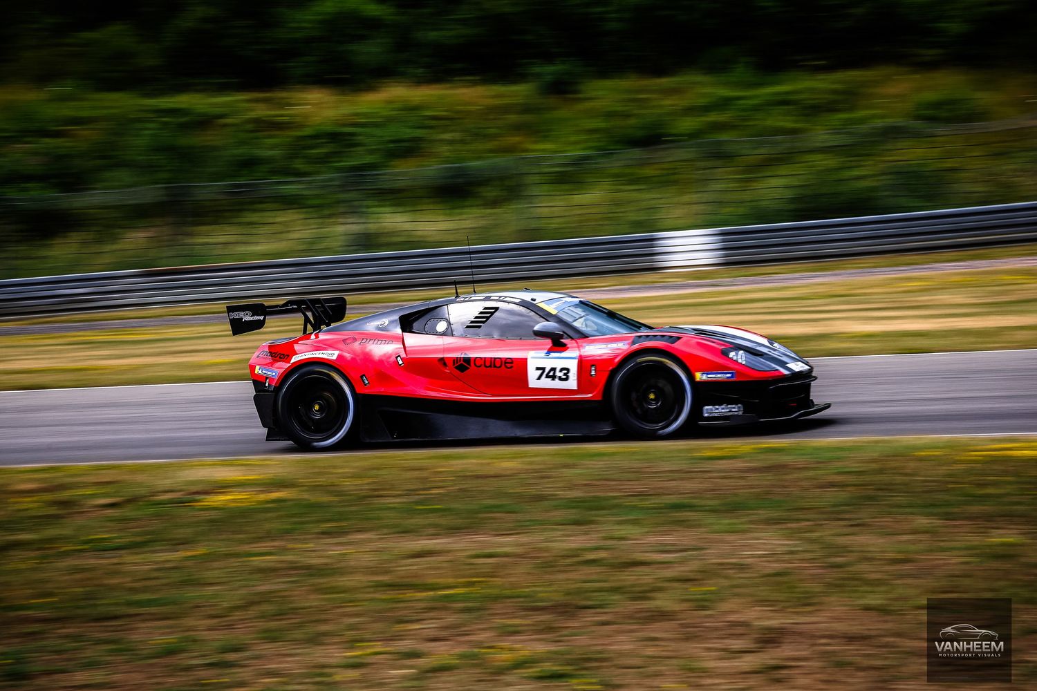 Red sports car number 743 racing on a track, captured with motion blur against a green landscape backdrop.