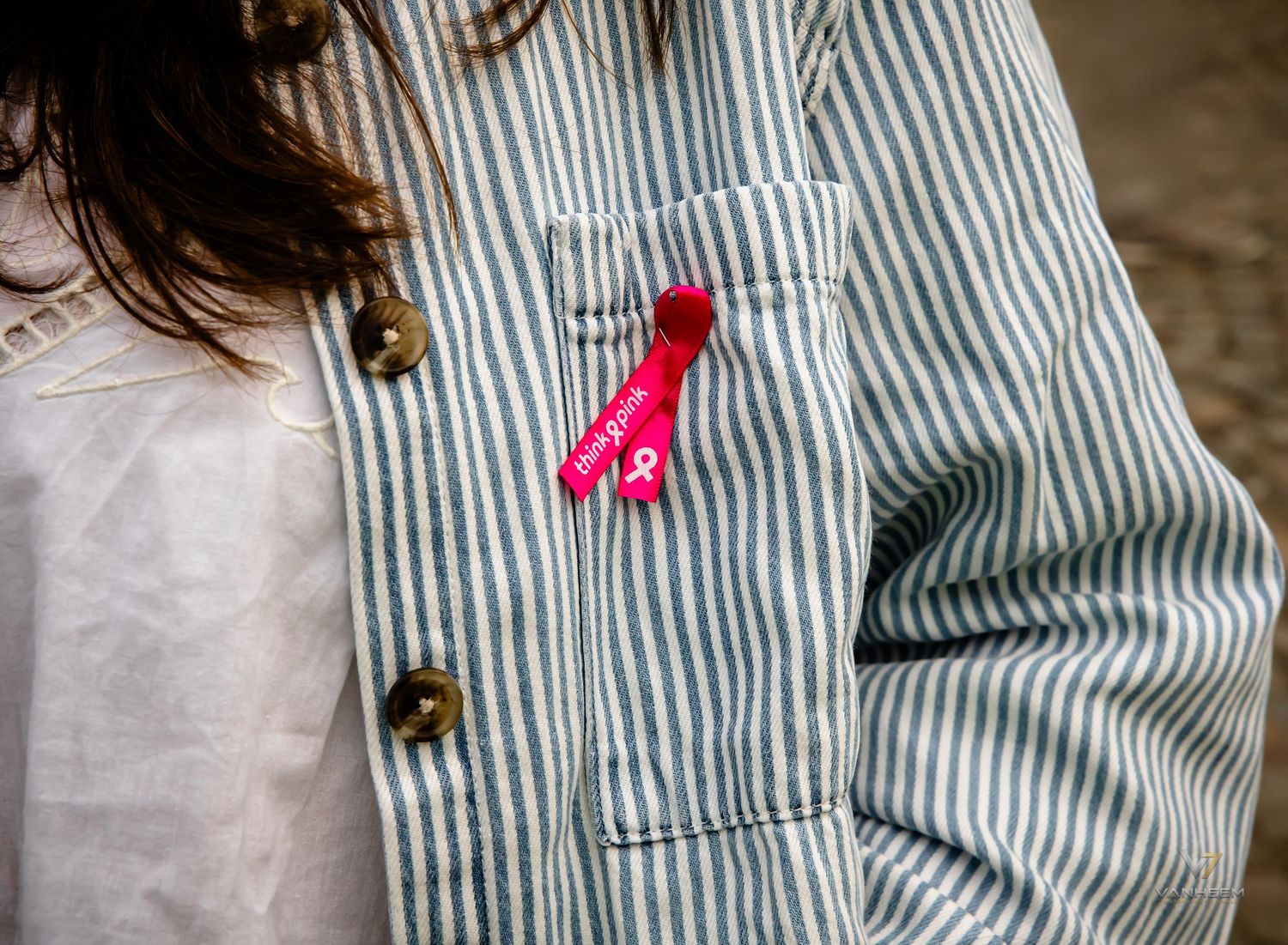 Close-up of a striped denim jacket with a pink ribbon breast cancer awareness pin on the chest pocket.