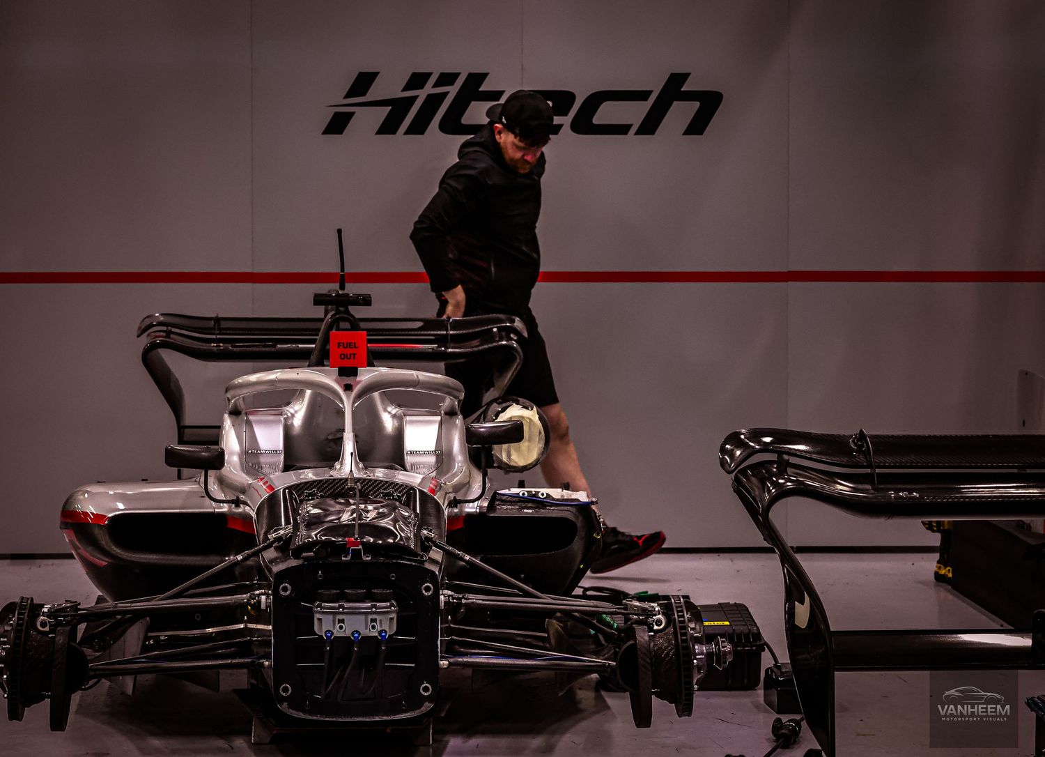 Mechanic working on a Hitech racing car in a dark pit garage during a motorsport event.
