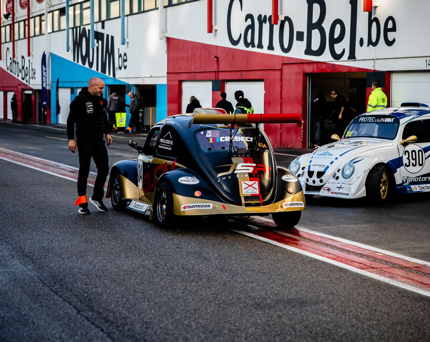 Modified VW Beetle race cars on pit lane at Carro-Bel.be circuit with mechanics during a motorsport event.