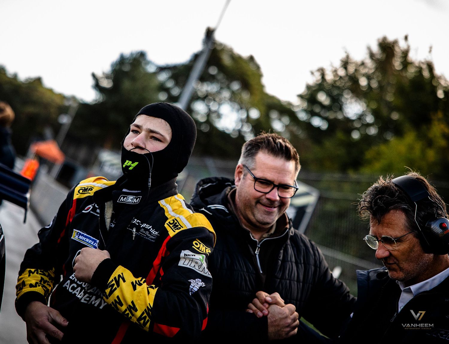Racing driver in black suit celebrates with team members at a motorsport event on a cloudy day.