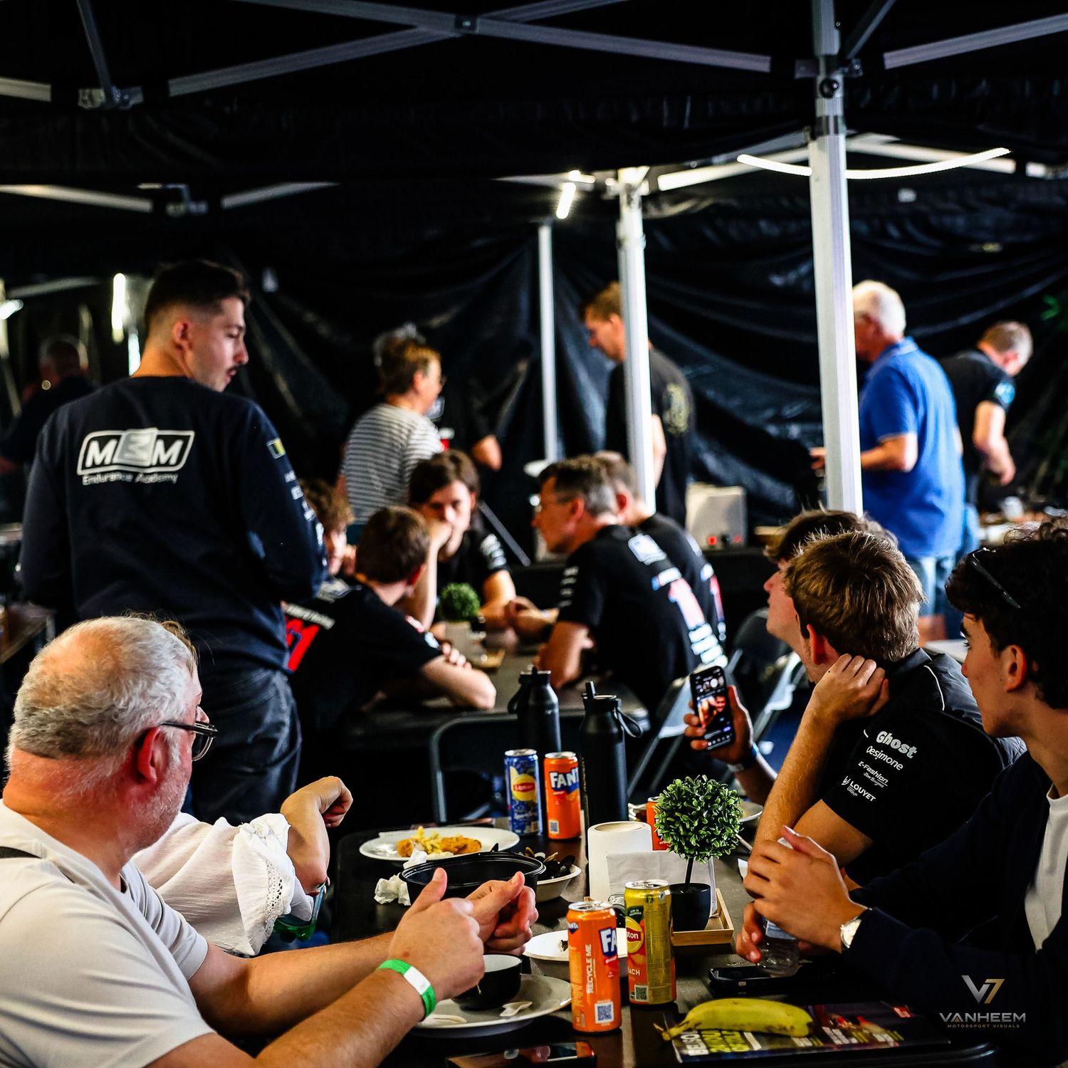 Racing team members gather under a tent at a motorsport event, dining together at tables with drinks and food.