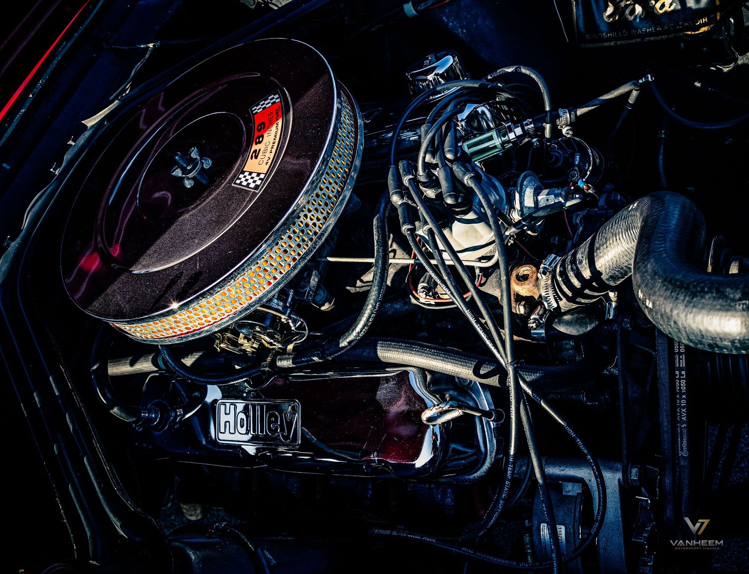 Close-up of a classic car engine bay featuring chrome components, air filter, and intricate wiring under dramatic lighting.