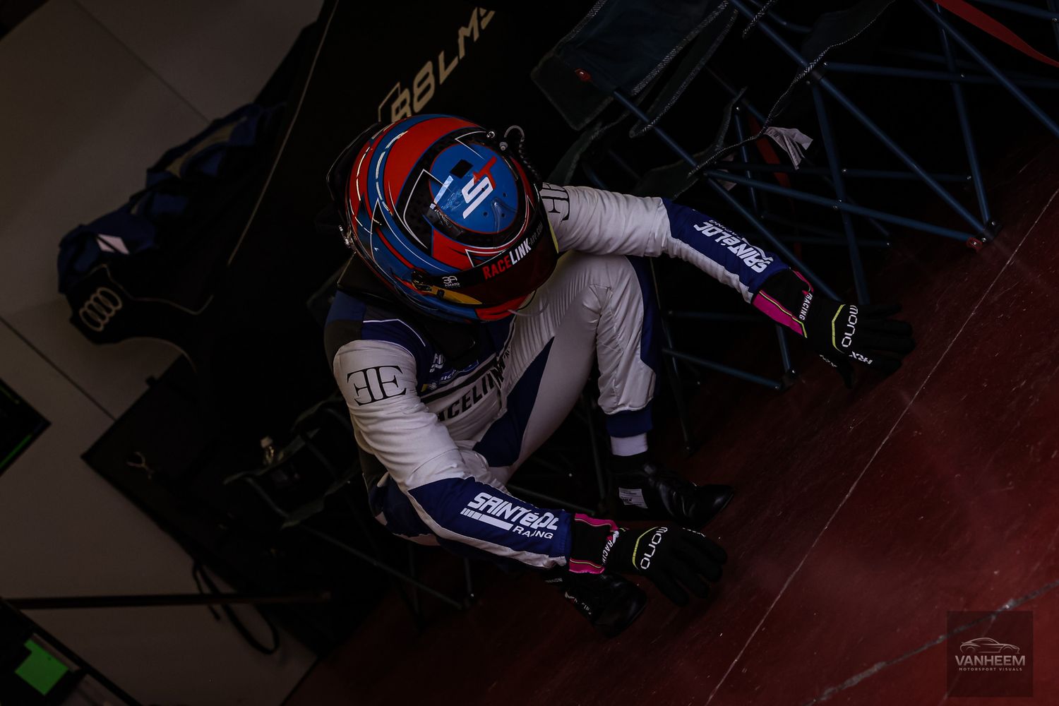A racing driver in white suit and blue helmet rests in a dark corner of what appears to be a pit area.