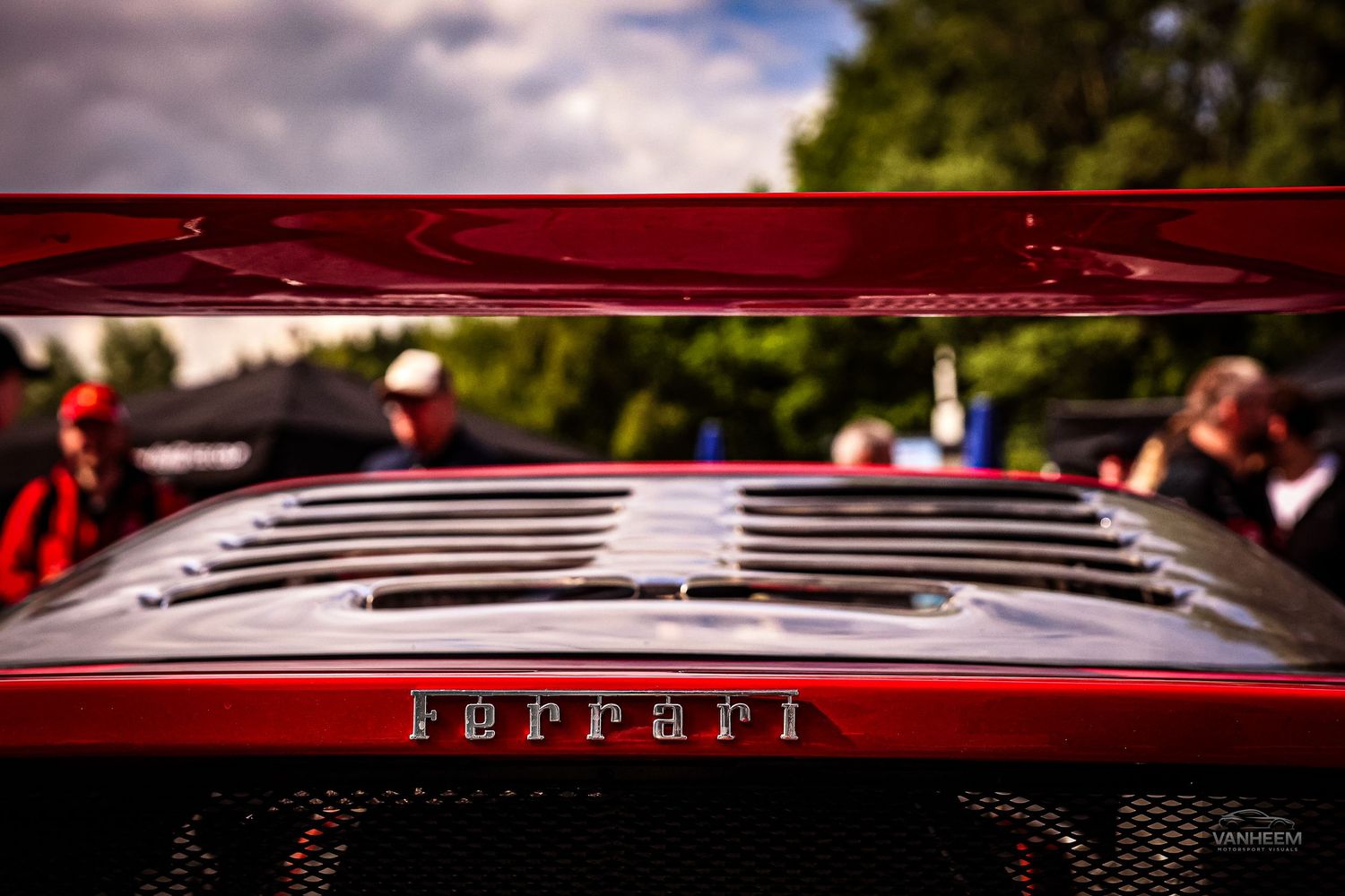 Close-up of a red Ferrari car hood with chrome badge, shallow depth of field, at an outdoor car show.