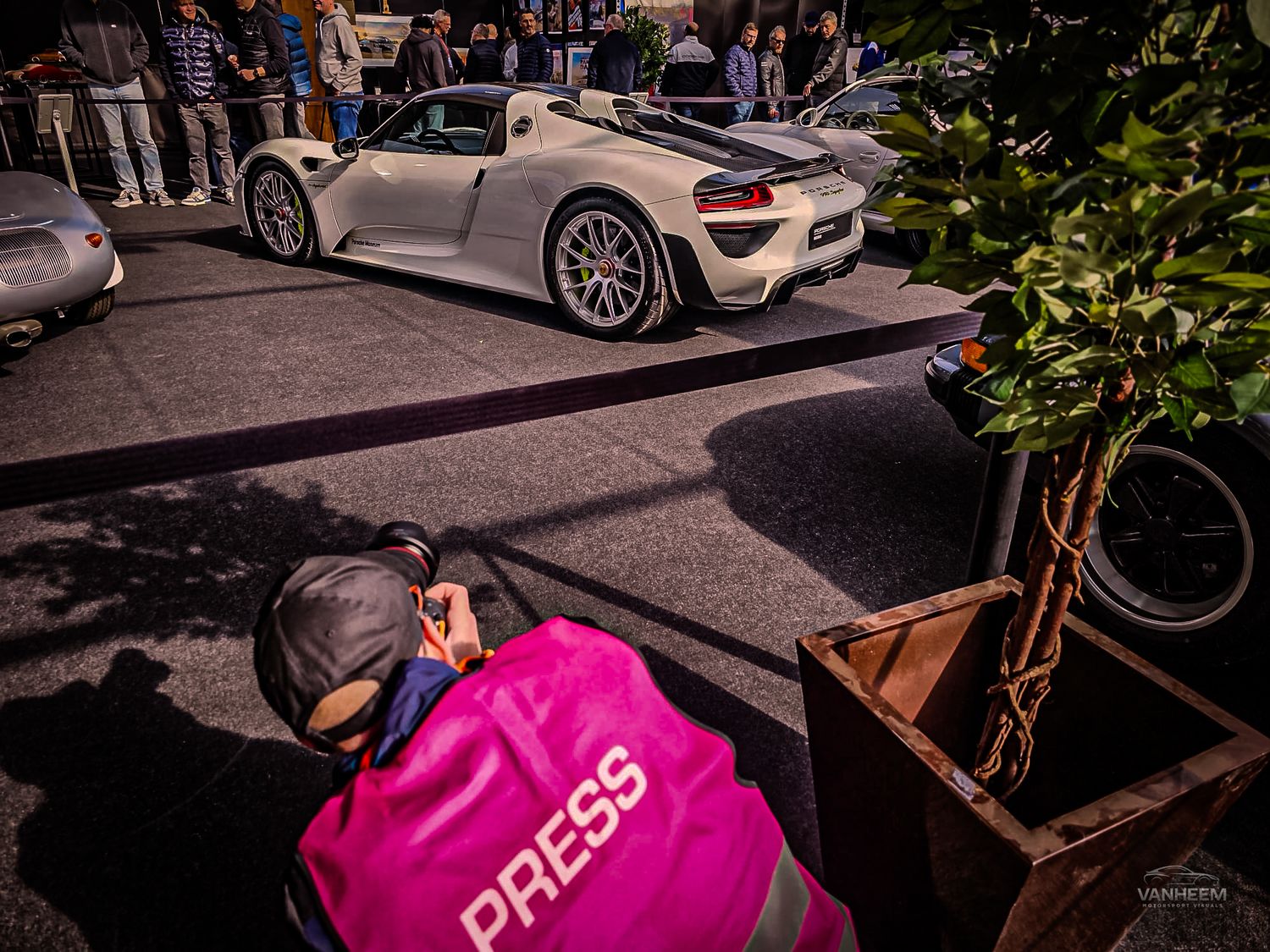 A press photographer in a pink jacket captures a white Porsche 918 Spyder at an outdoor automotive event.