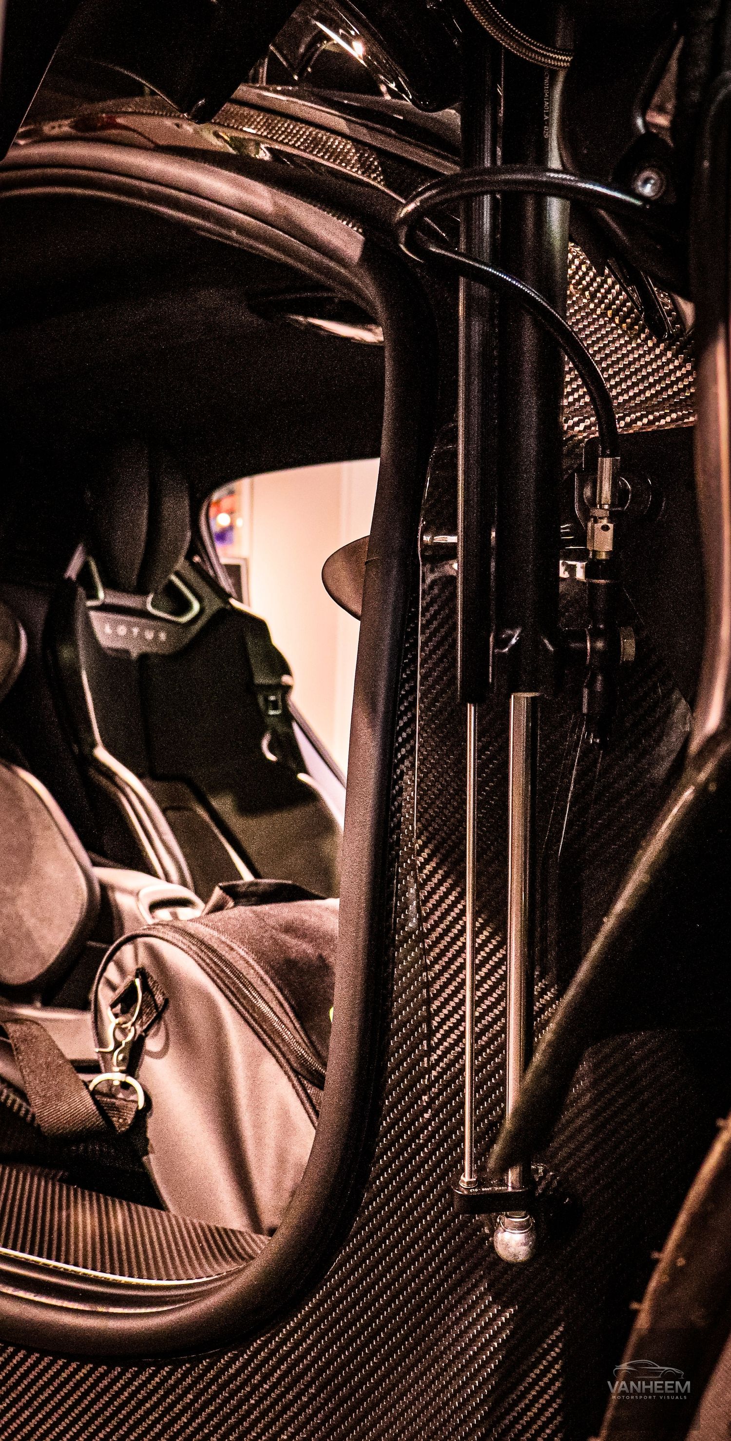 Close-up of a Lotus supercar carbon fiber interior showing racing seat, roll cage, and suspension components.