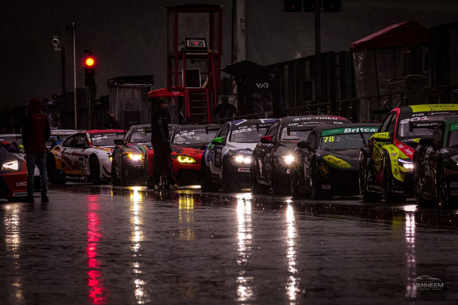 Race cars lined up in wet pit lane at night, headlights reflecting on rain-soaked track during motorsport event.