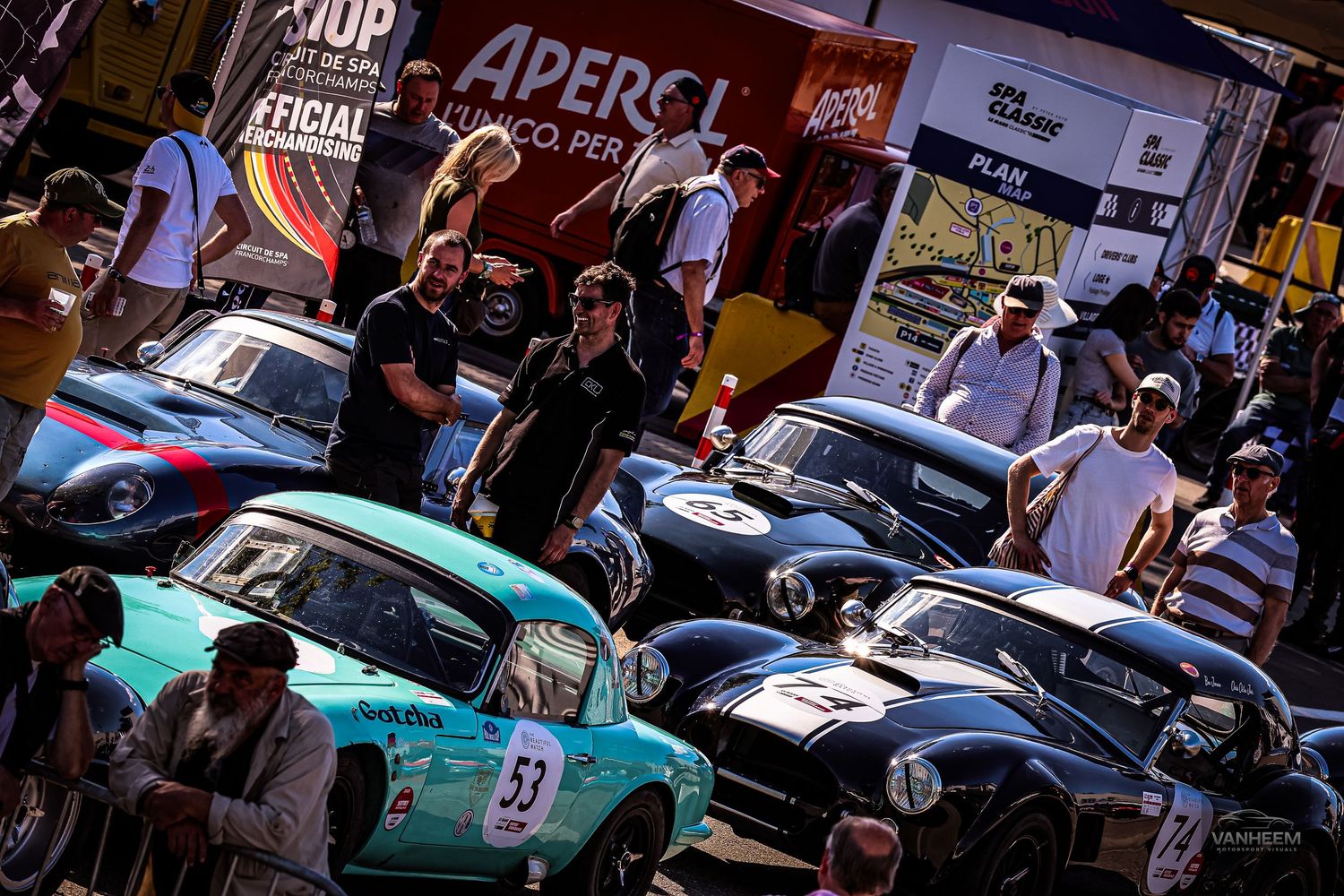 Vintage racing cars lined up in pit lane at a classic car race event with Aperol sponsorship banners and spectators.