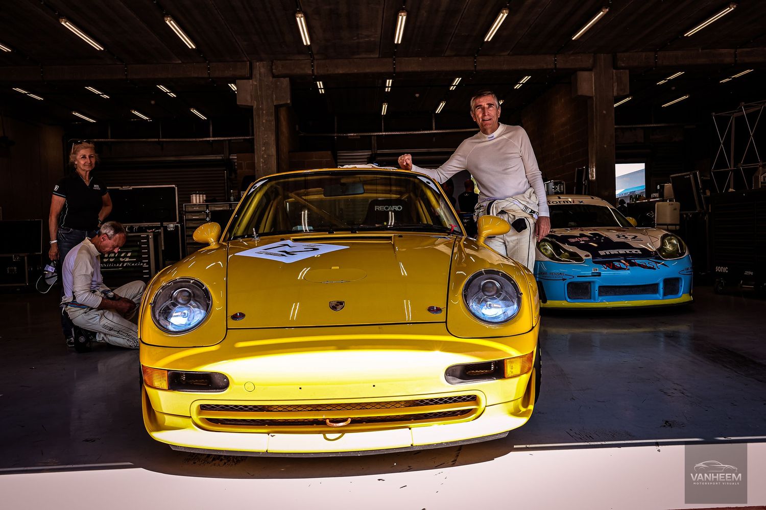 Yellow Porsche 993 race car in a garage with mechanics and a blue Porsche in the background.