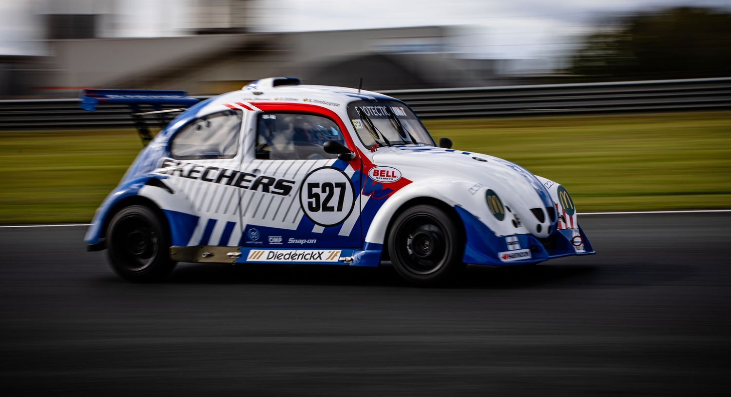 A Skechers-sponsored VW Beetle race car numbered 527 speeds around a motorsport circuit track.