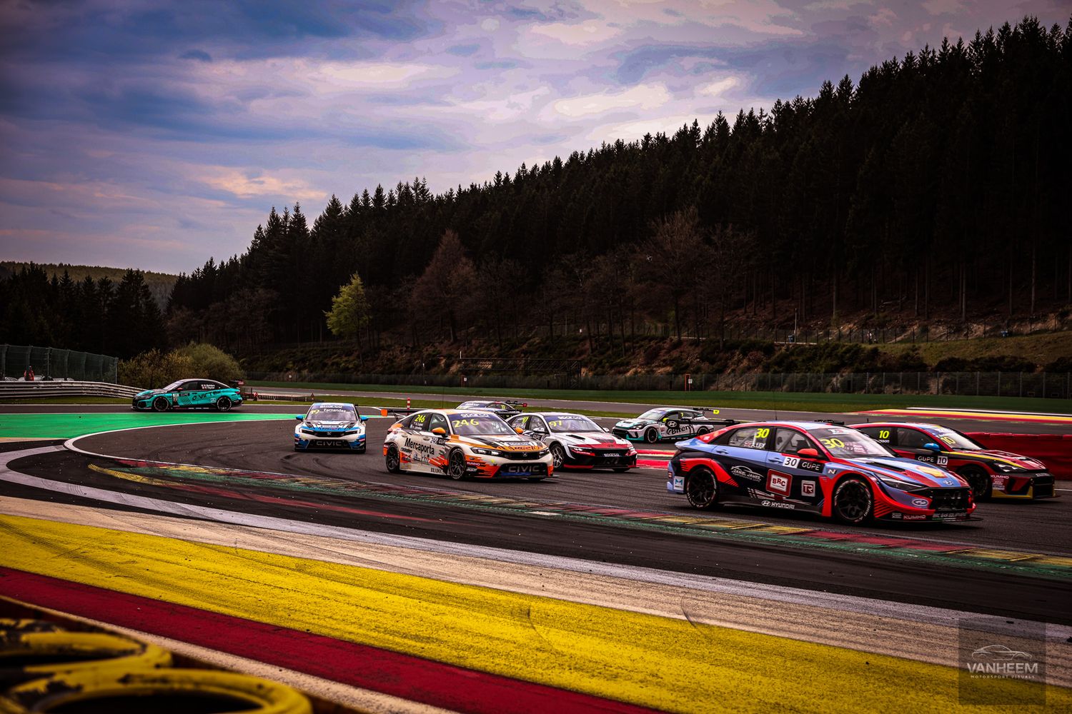 Multiple racing cars compete on a winding circuit track at Spa-Francorchamps during an exciting motorsport event.