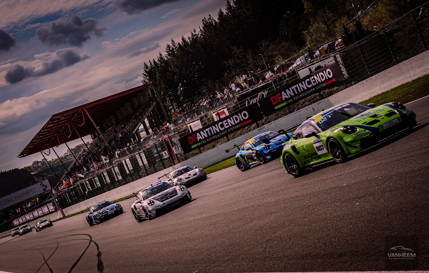 Green race car leads a pack of GT cars on a circuit track during a cloudy motorsport racing event.