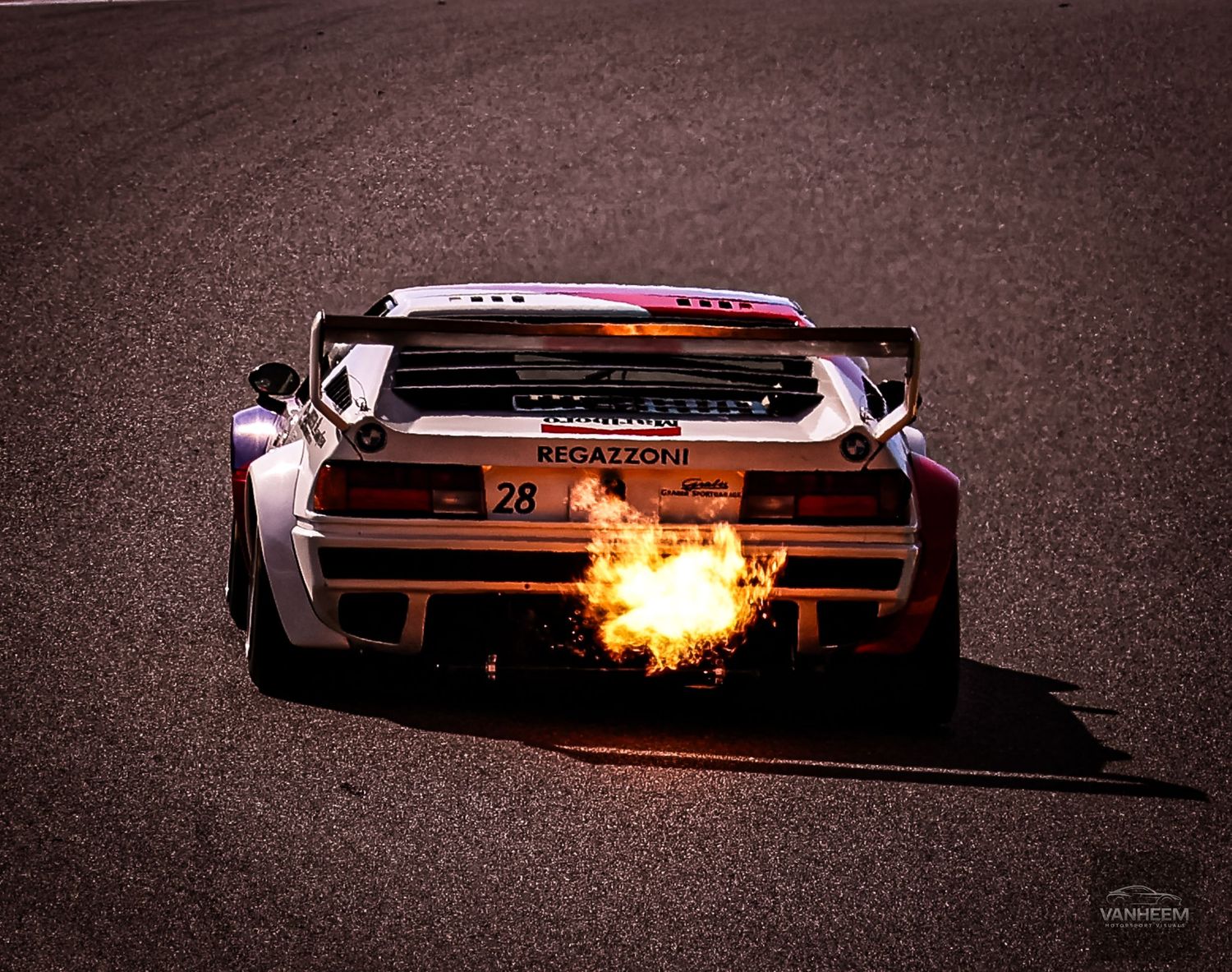 Race car number 28 spitting flames from exhaust on track during high-speed motorsport competition event.