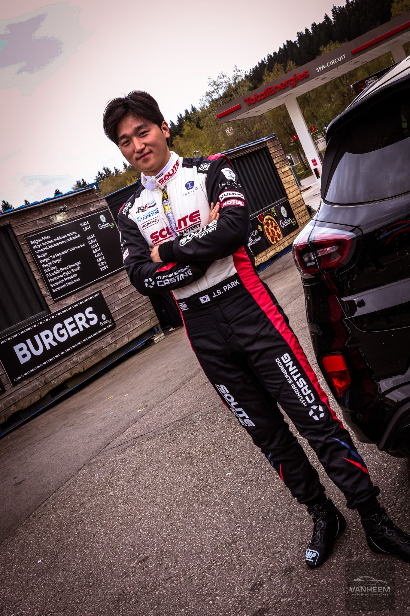 Racing driver in black sponsor suit leans against a car at Spa Circuit near a burgers sign.