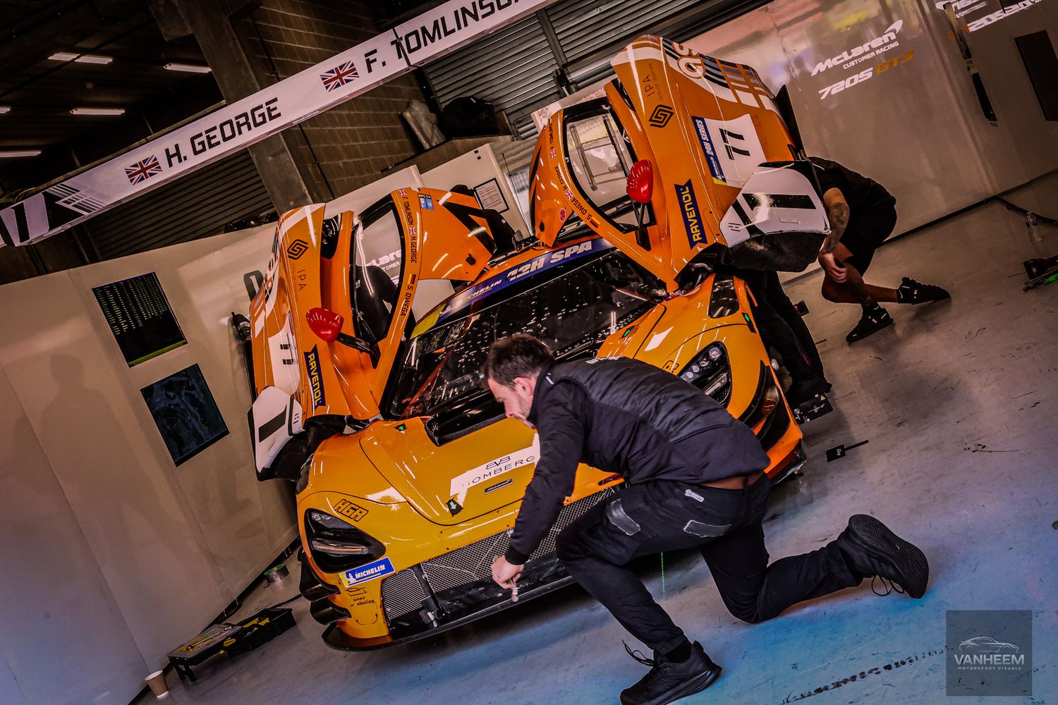 McLaren GT racing car in orange livery being serviced by mechanics in a pit garage during a motorsport event.