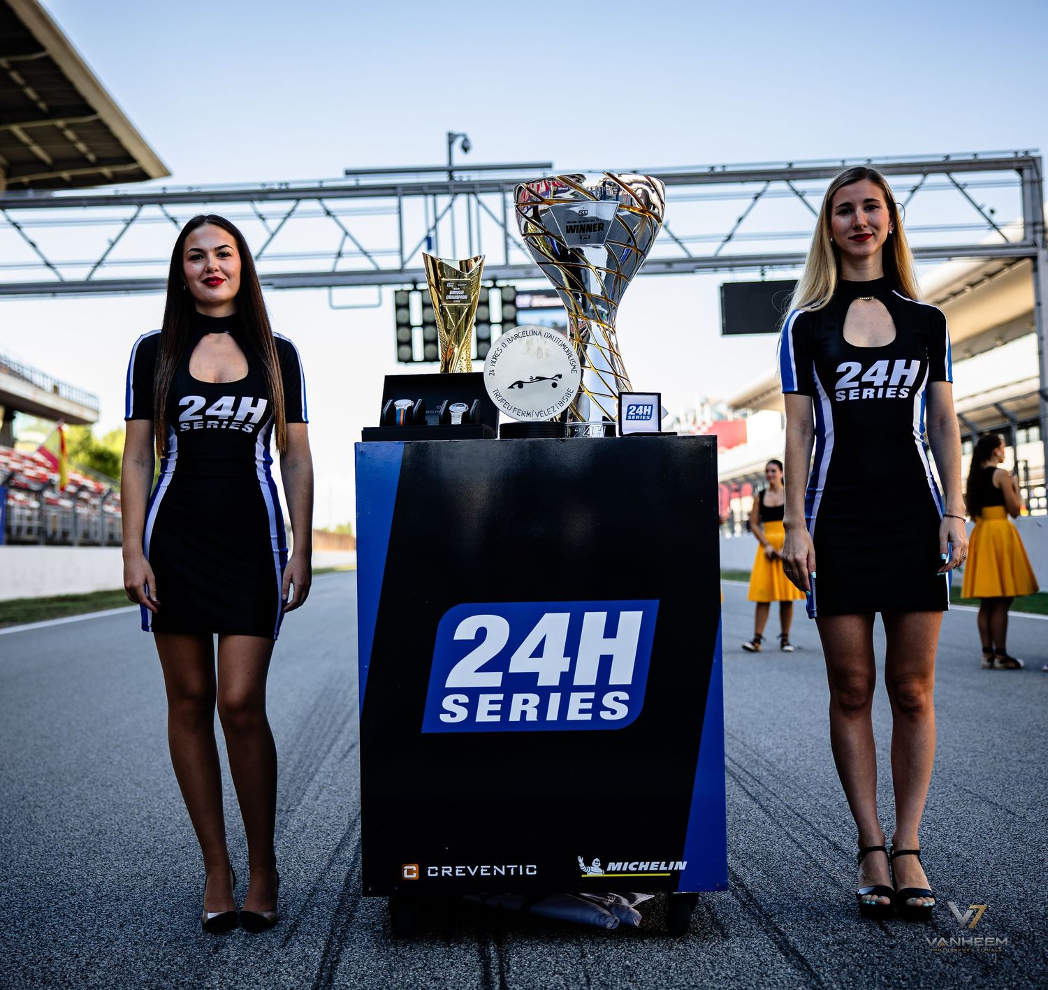 Two grid girls in black uniforms flank a trophy display for the 24H Series endurance racing event.