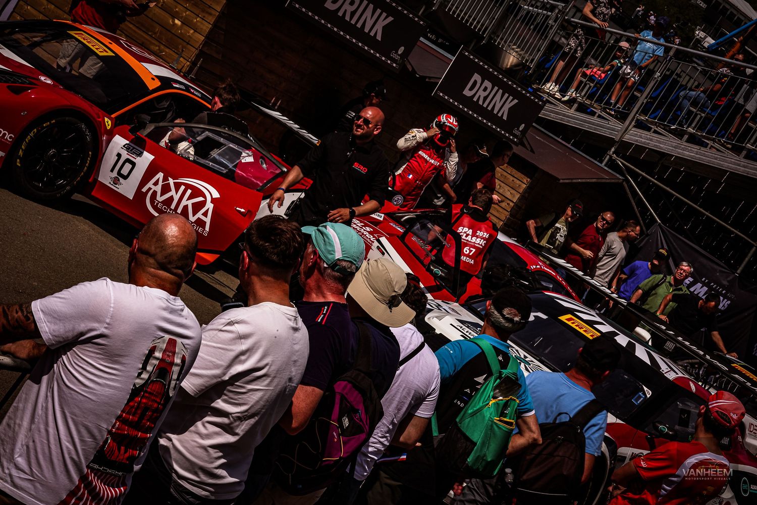 Crowd of racing fans gather around red IMSA race cars in a busy pit lane area during a motorsport event.
