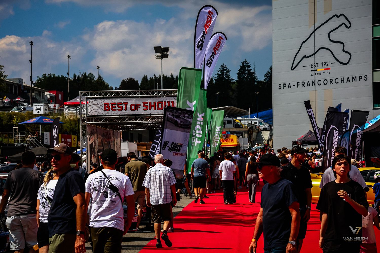 Crowds walk along a red carpet at Spa-Francorchamps racing circuit event with colorful sponsor flags and Best of Show signage.