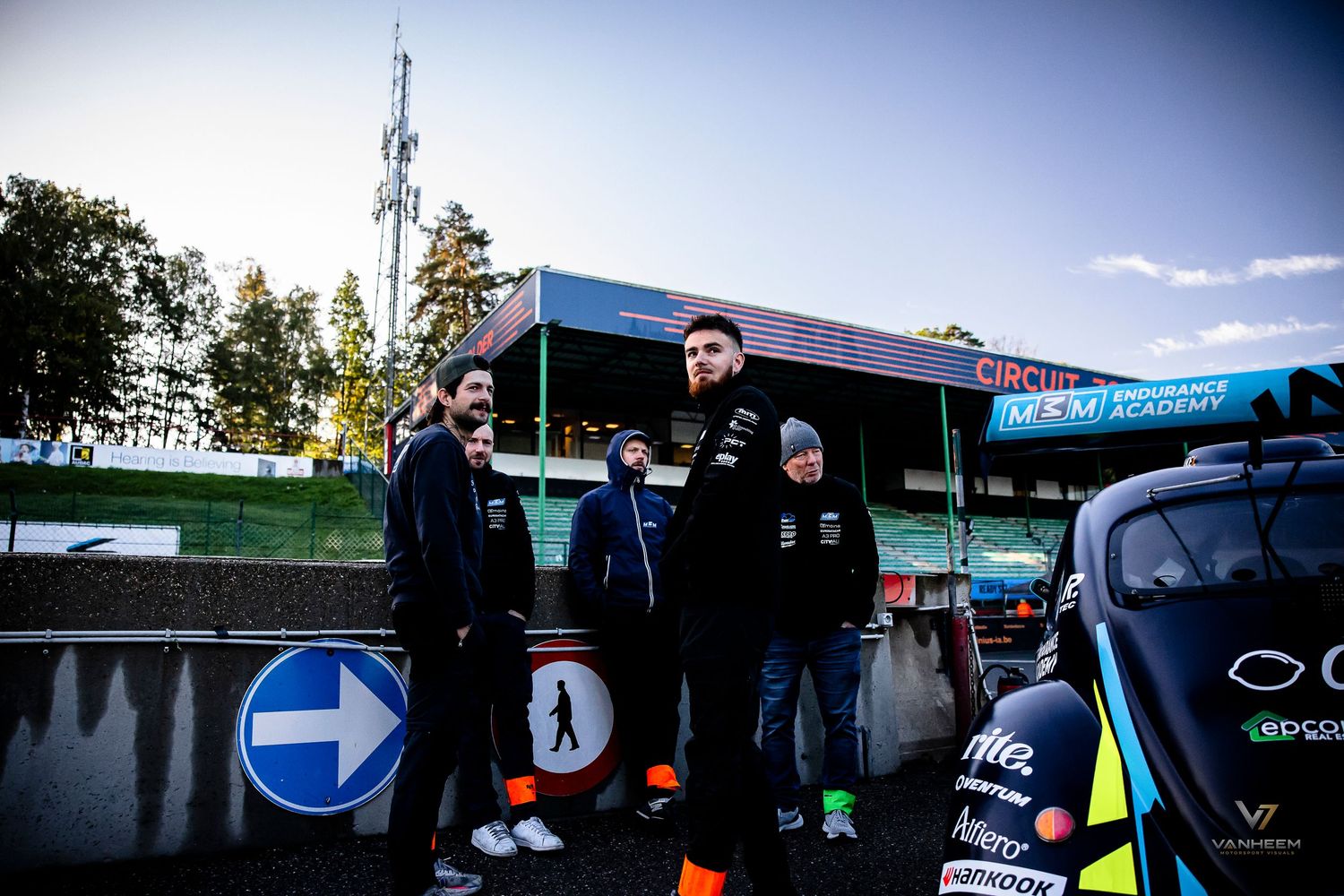 Racing team members stand near a race track pit area with directional signs and a race car visible.