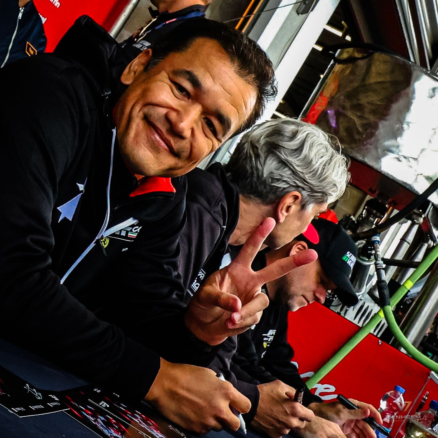 Two men smiling and making peace signs while signing autographs at a racing event.