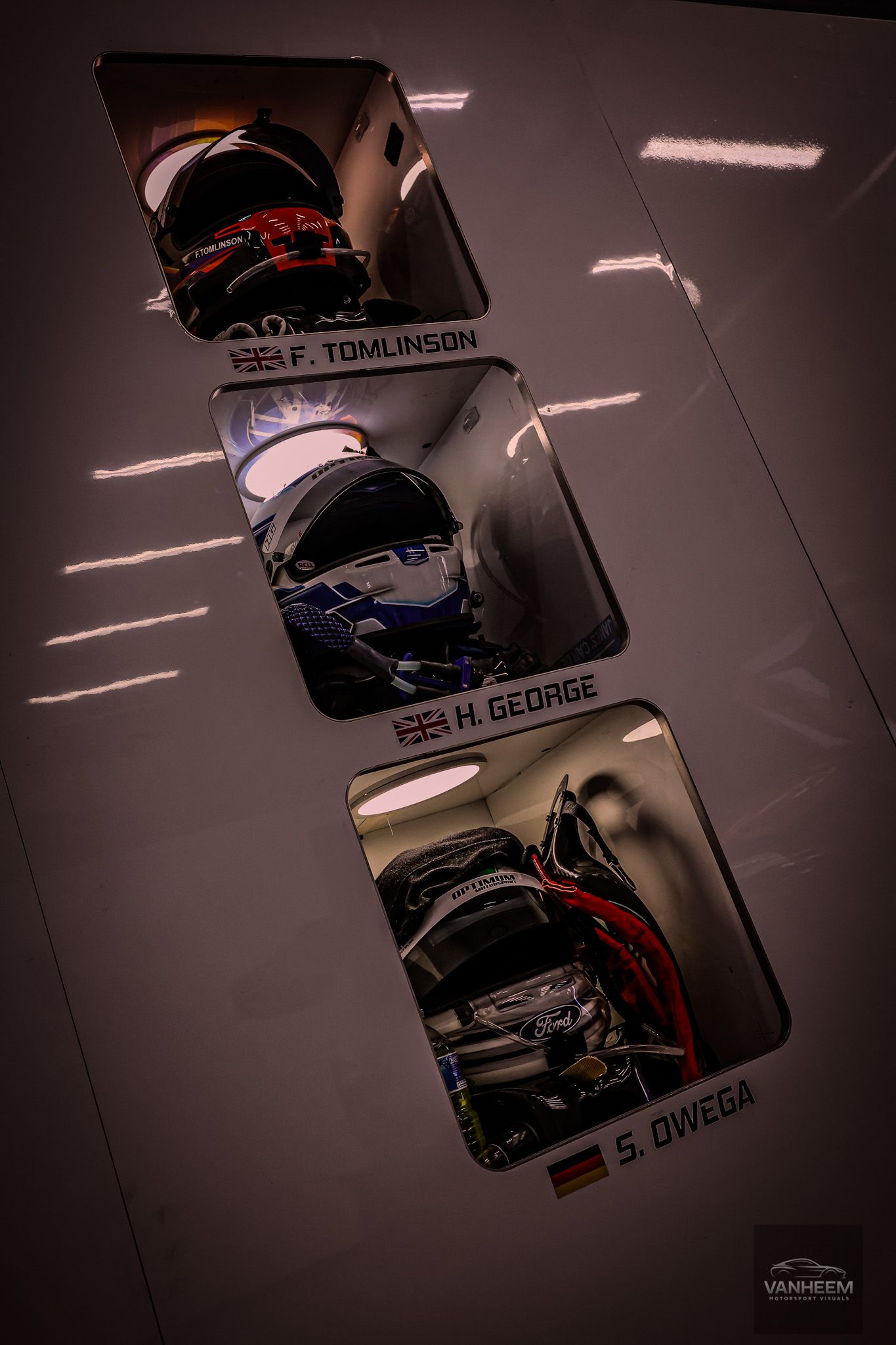 Three racing helmets displayed in illuminated wall-mounted cases labeled F. Tomlinson, H. George, and S. Owega at a motorsport event.