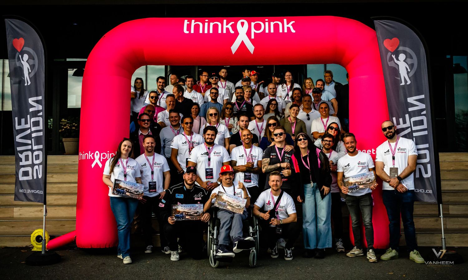 Large group poses under pink Think Pink arch at breast cancer awareness charity event outdoors.