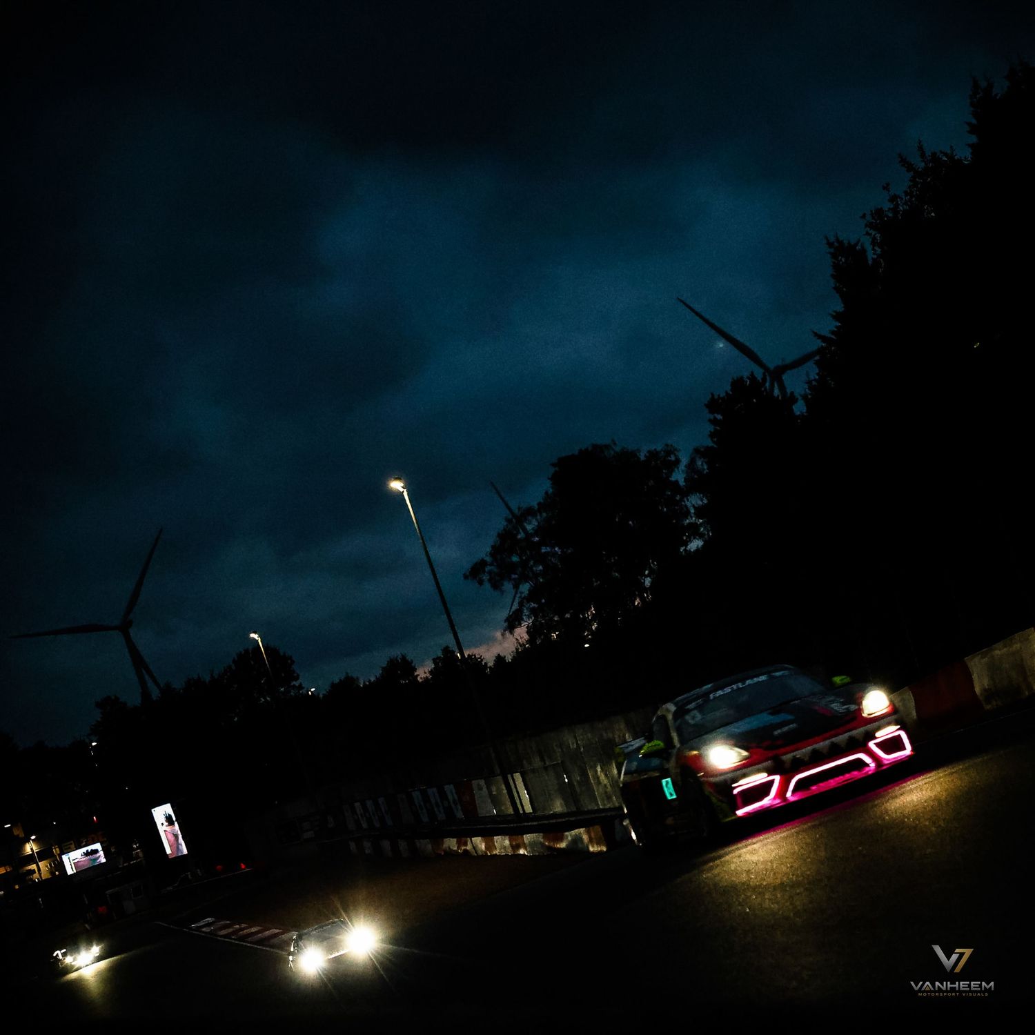 A racing car with glowing pink lights speeds through a dark circuit at night under dramatic stormy skies.