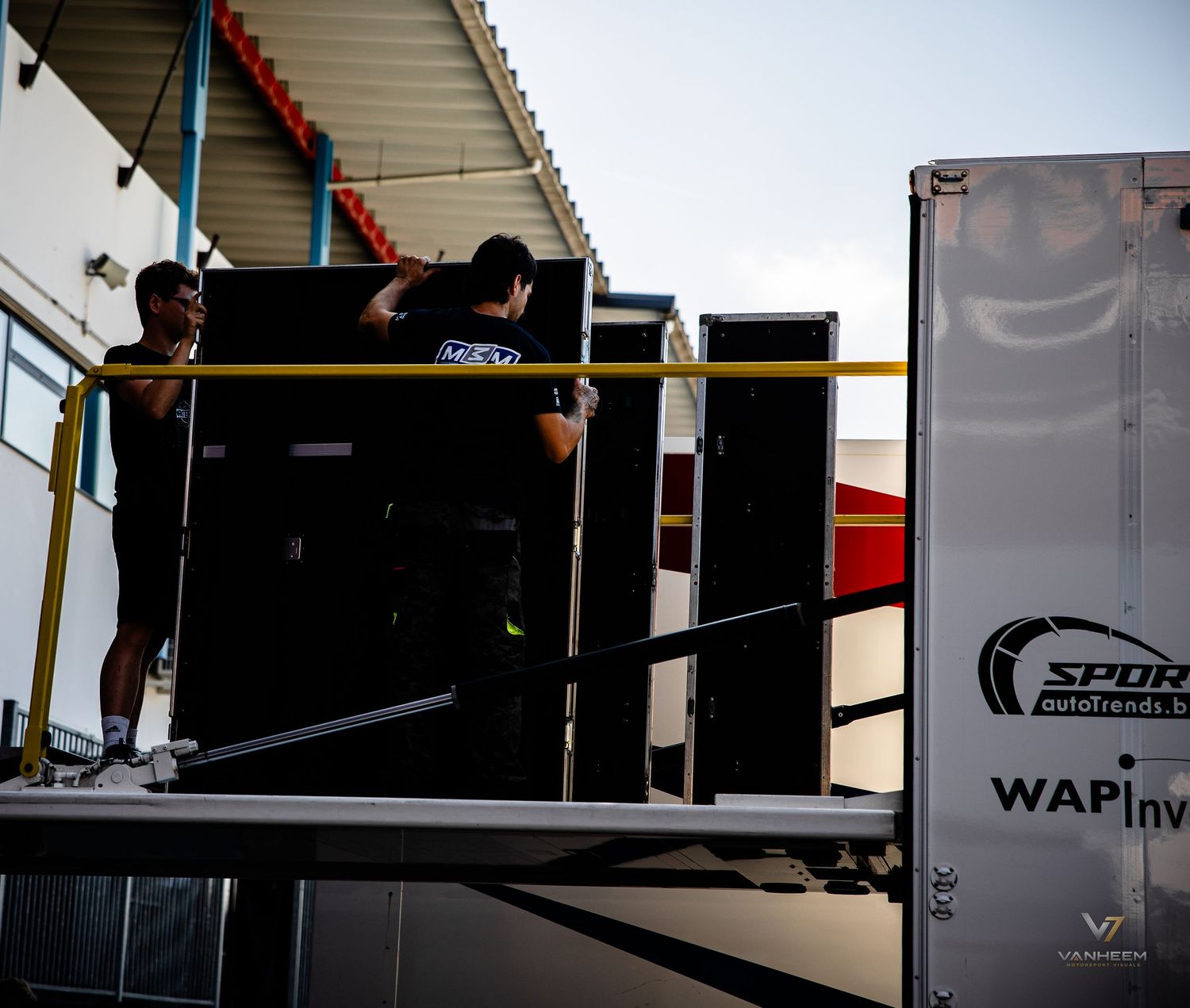 Two workers unloading large equipment from a Sport Automobiles branded trailer at a racing event venue.