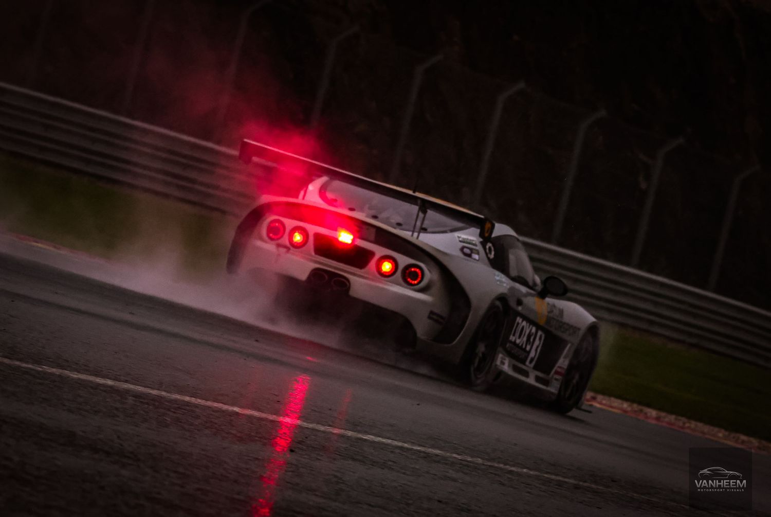 A racing car speeds on a wet track at night, its red brake lights glowing through the misty rain and spray.