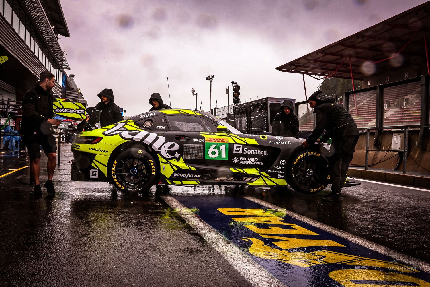 Race car #61 in yellow-green livery during a wet pit stop at a motorsport event with crew members working in rain.