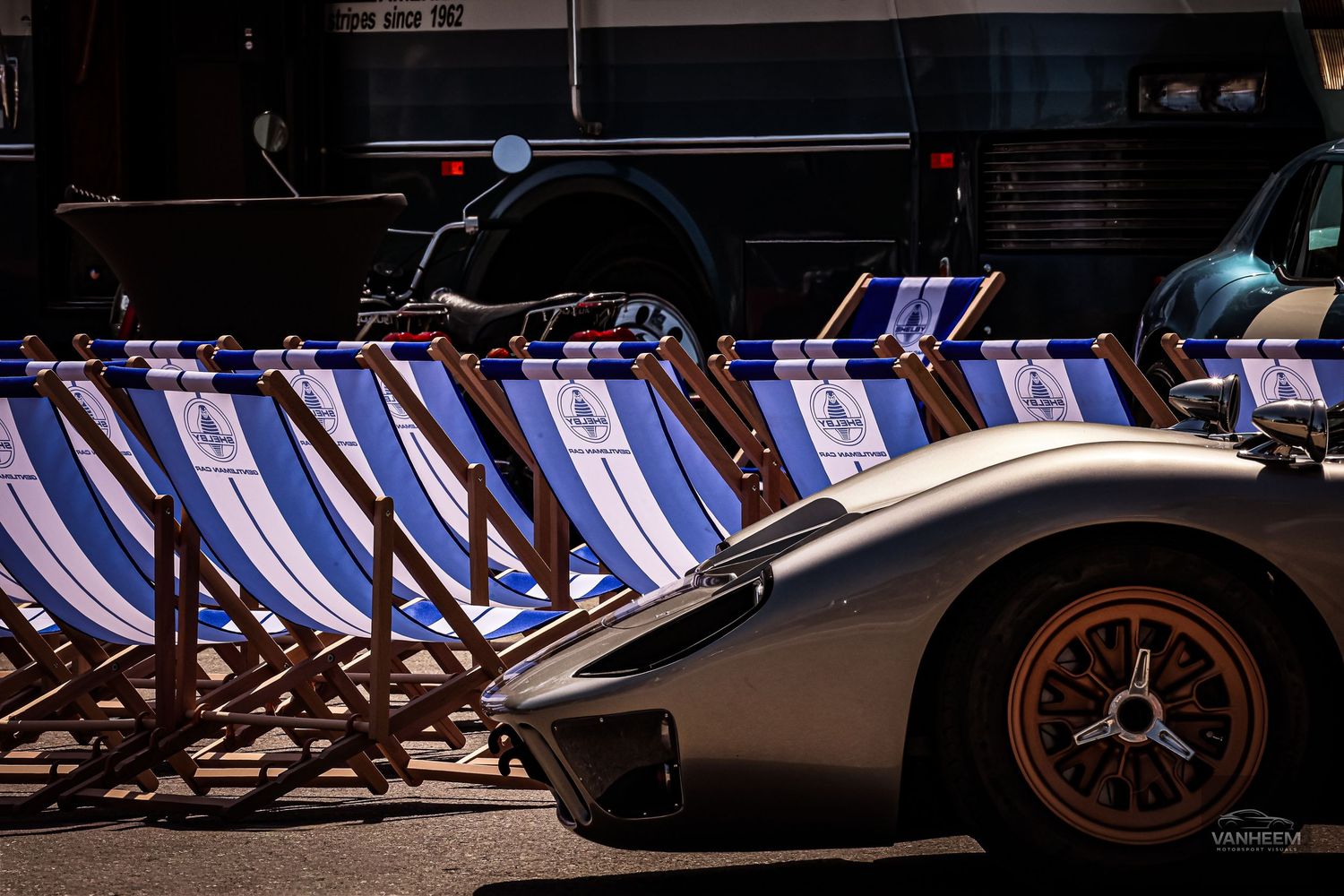 Silver sports car parked beside a row of blue and white striped deck chairs at a sunny outdoor event.