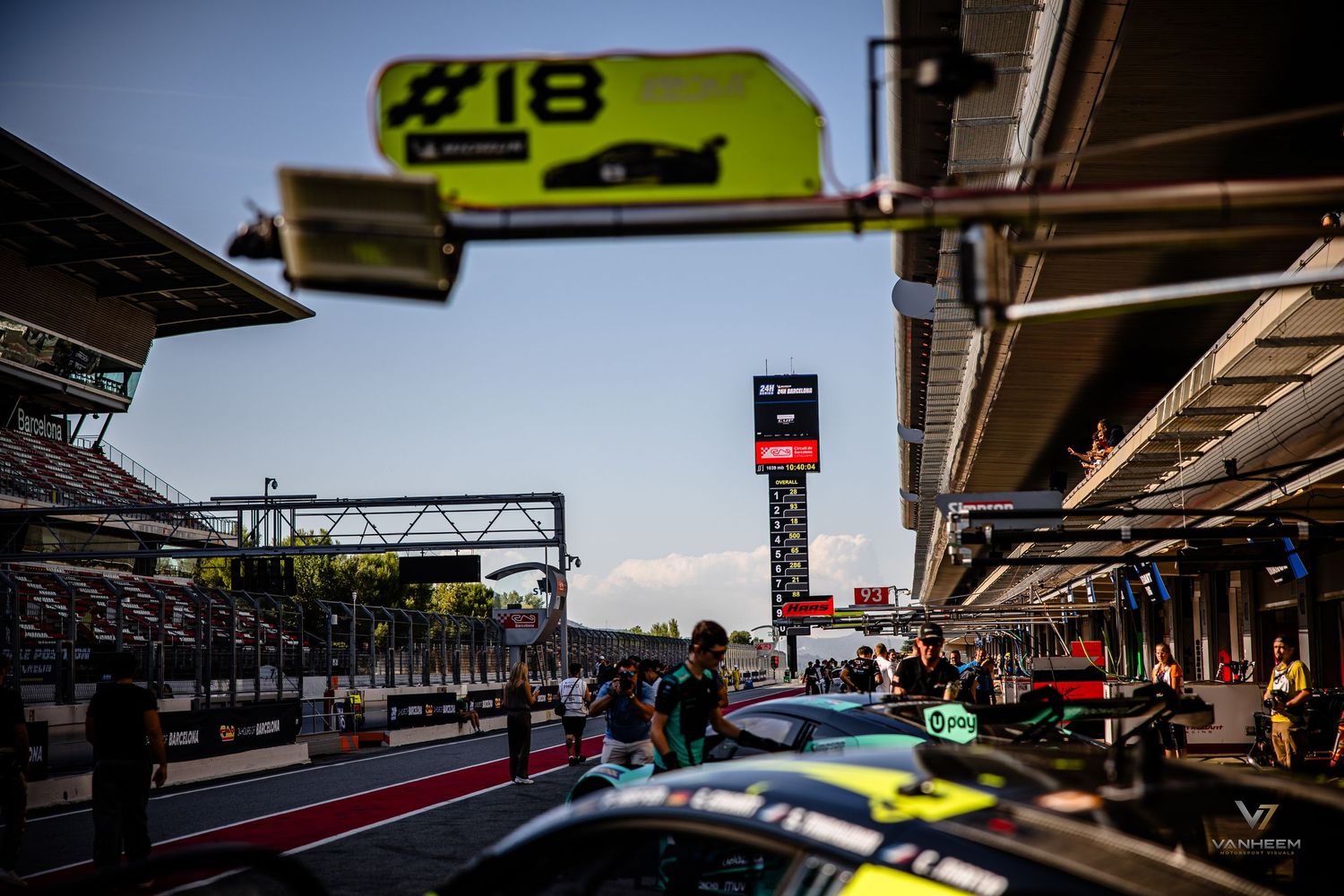 Racing cars line up in pit lane at a motorsport circuit under a number 18 pit board sign during a race event.