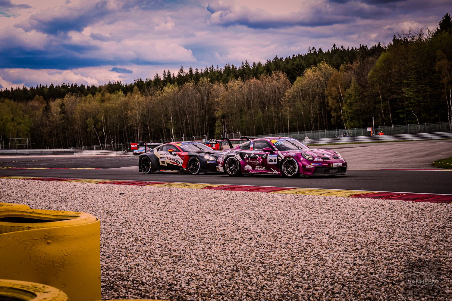Two pink livery GT racing cars battling side by side on Spa-Francorchamps circuit under dramatic cloudy sky.