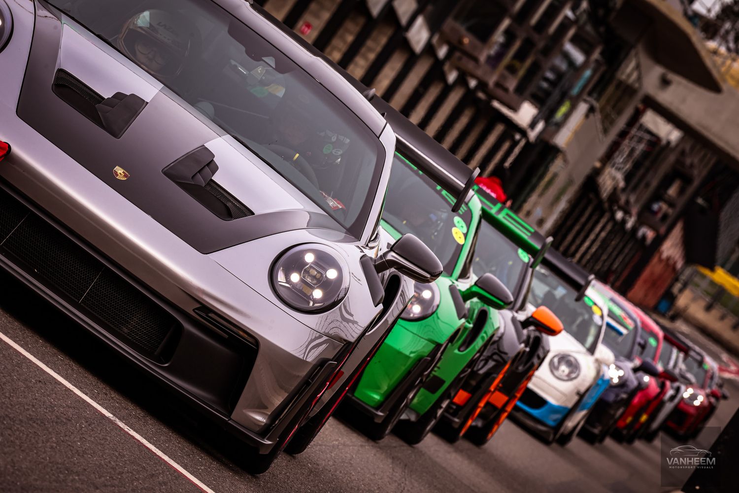 A colorful lineup of Porsche sports cars parked on a city street, showcasing various models in silver, green, and multicolor liveries.
