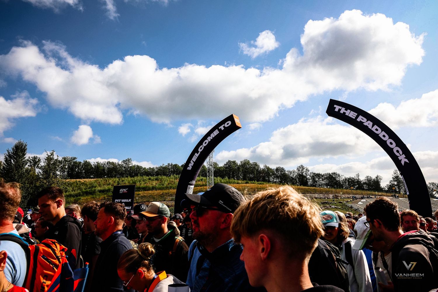 Crowd of fans entering The Paddock arch at an outdoor motorsport event under a partly cloudy sky.