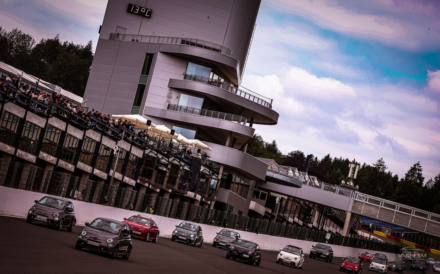 Multiple racing cars compete on a circuit track with a white control tower and spectators in the background.