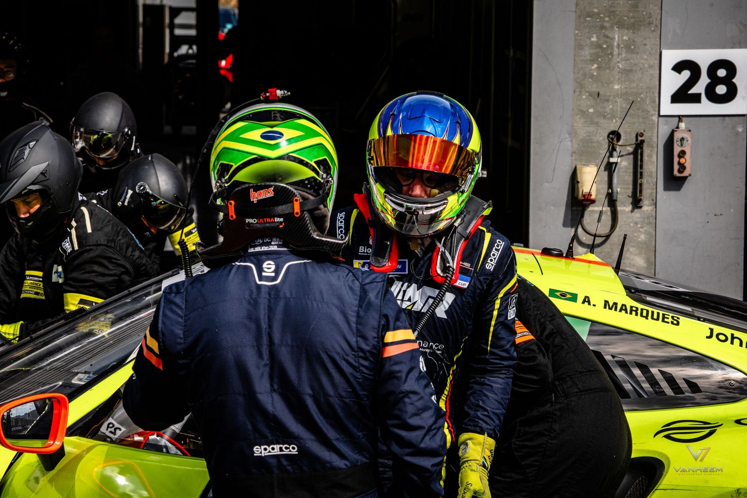 Two racing drivers in colorful helmets embrace in the pit lane near car 28 during a motorsport event.