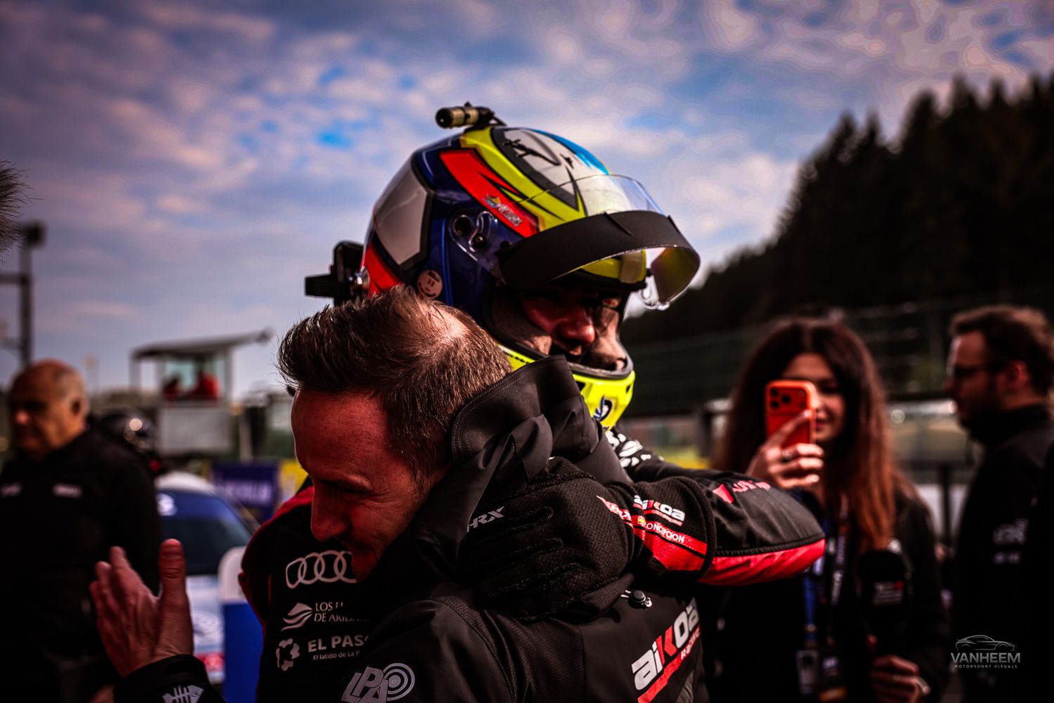 Racing driver in helmet celebrates with team members at motorsport event during dusk at a racing circuit.