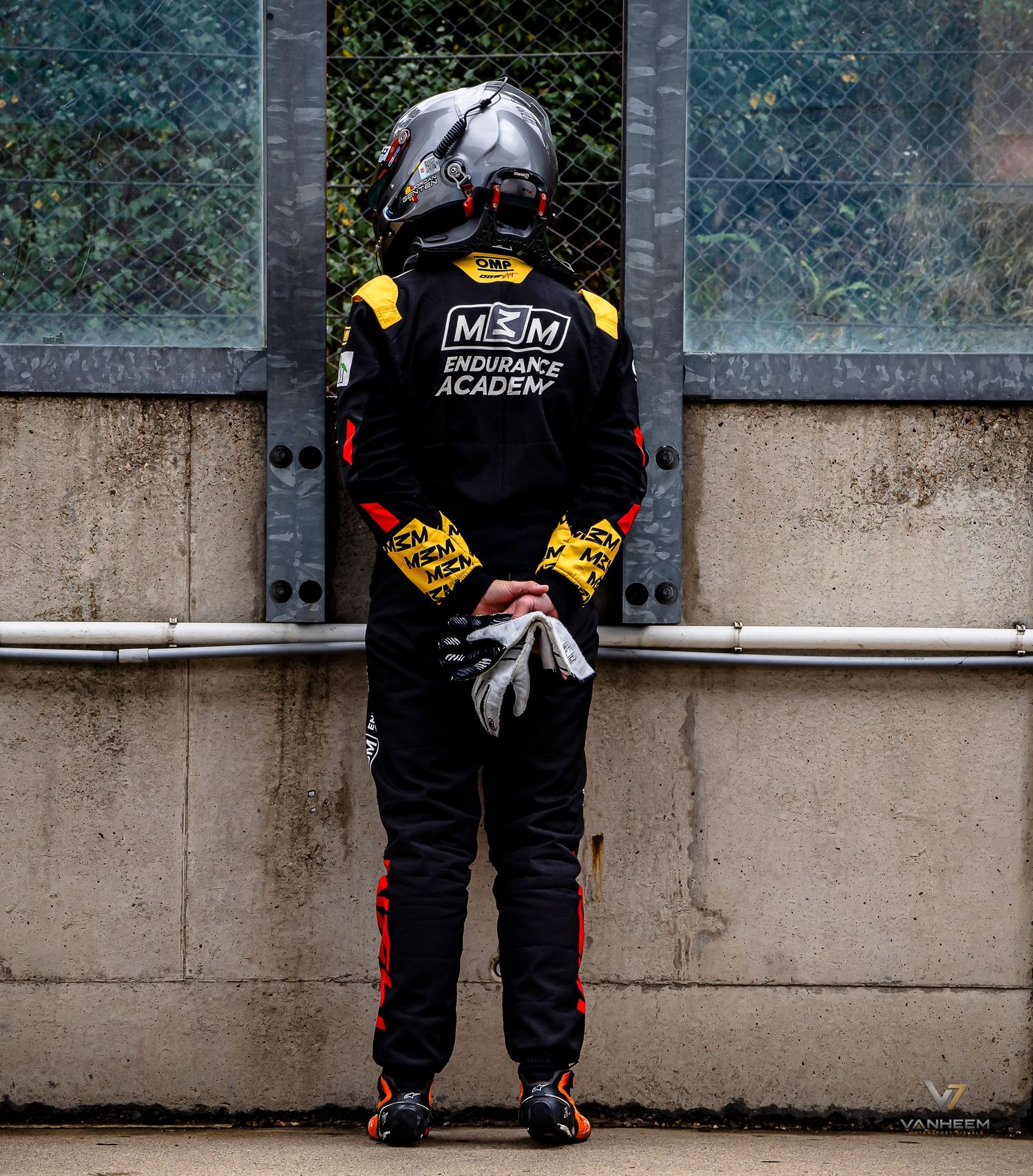 Racing driver in MCM Endurance Academy suit and helmet stands facing a wall, holding gloves behind their back.