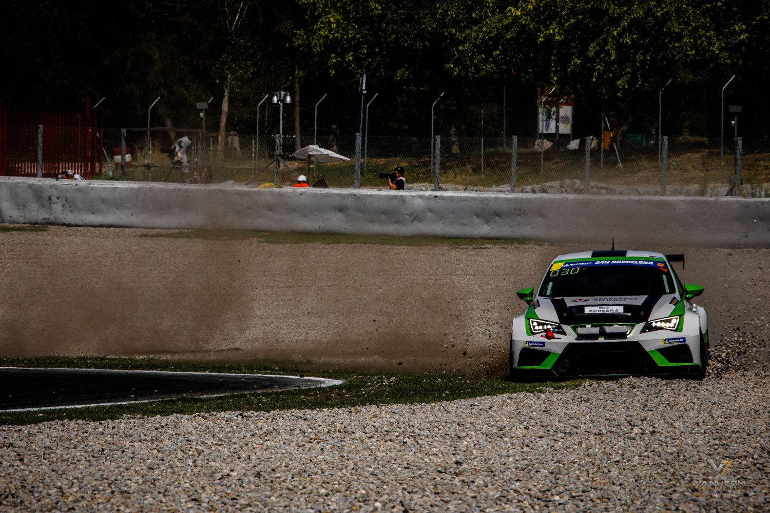 Green and white rally car racing on a circuit track with gravel runoff area and trees in the background.