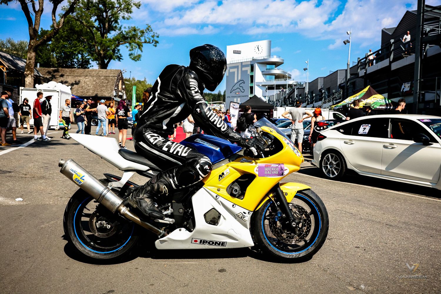 Rider in black leather gear on a yellow and white sport motorcycle at an outdoor motorsport event on a sunny day.