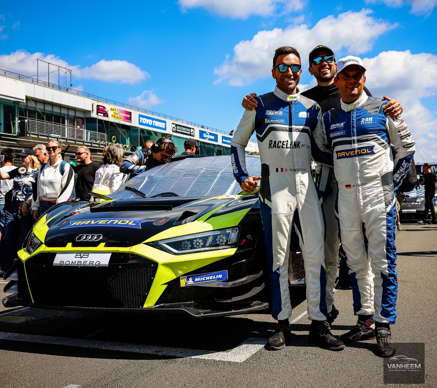 Three racing drivers pose beside an Audi GT3 race car at a motorsport event on a sunny day.