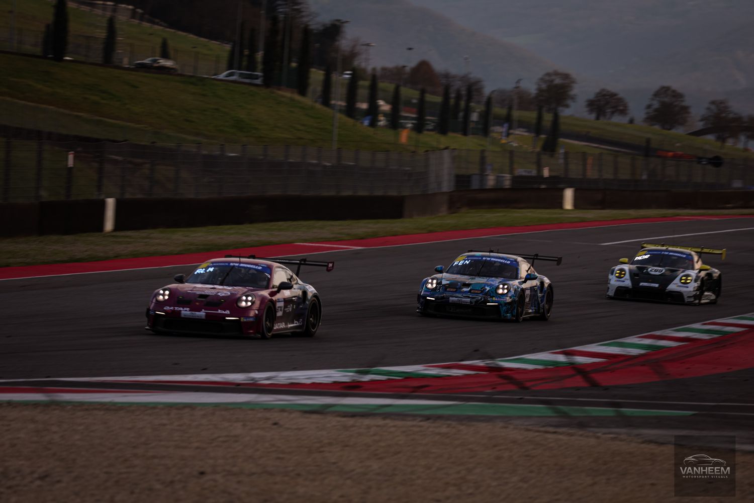 Three race cars navigate a curve on a professional racing circuit during dusk competition.
