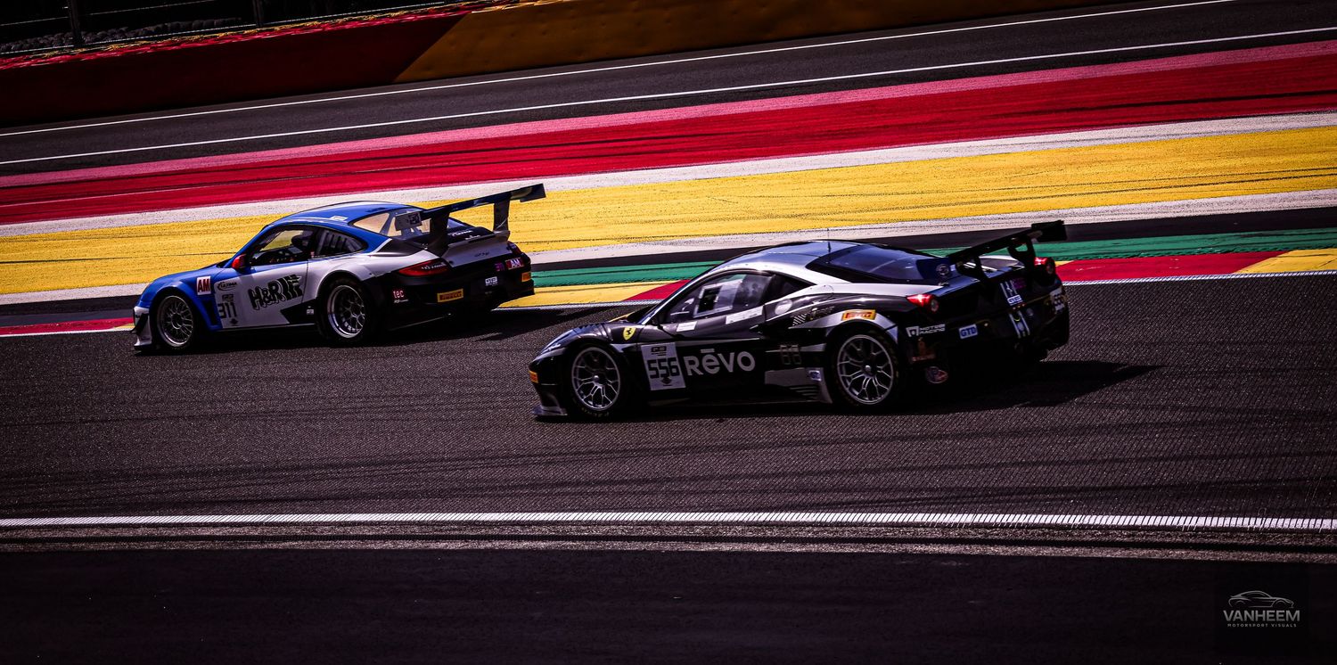 Two racing cars competing on a circuit track, featuring a black sports car leading a blue and red GT race car on asphalt.