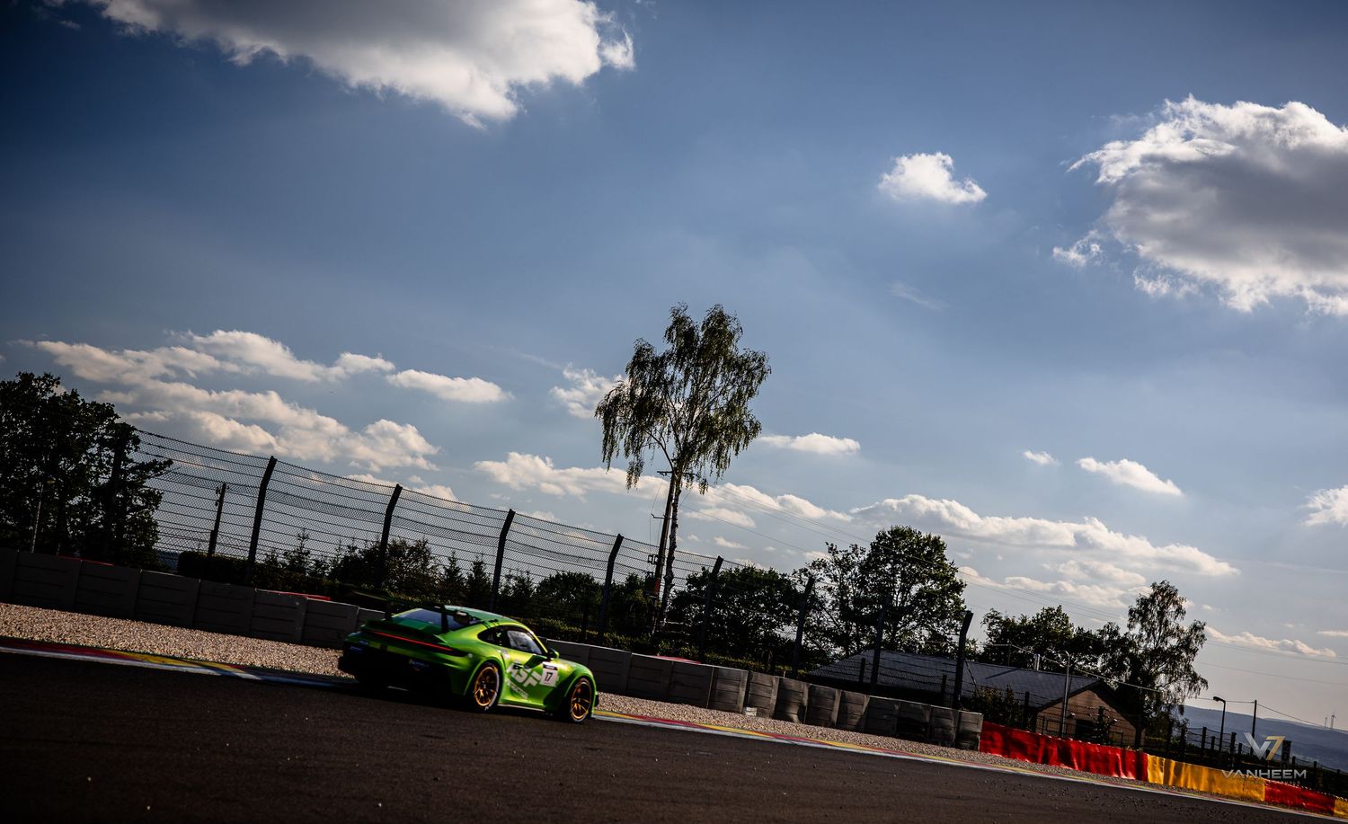 Green racing car speeding on a circuit track under a dramatic cloudy sky with trees in the background.