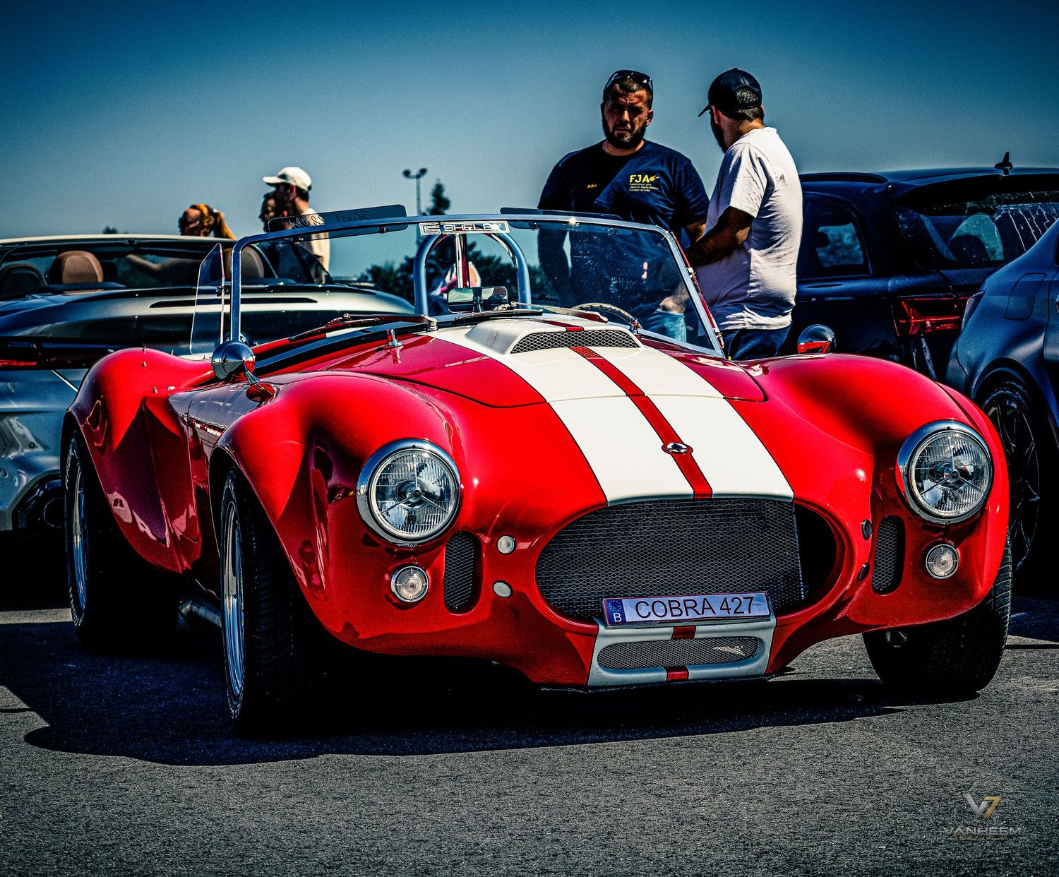 Classic red Shelby Cobra with white racing stripes at a car show, surrounded by spectators on a sunny day.