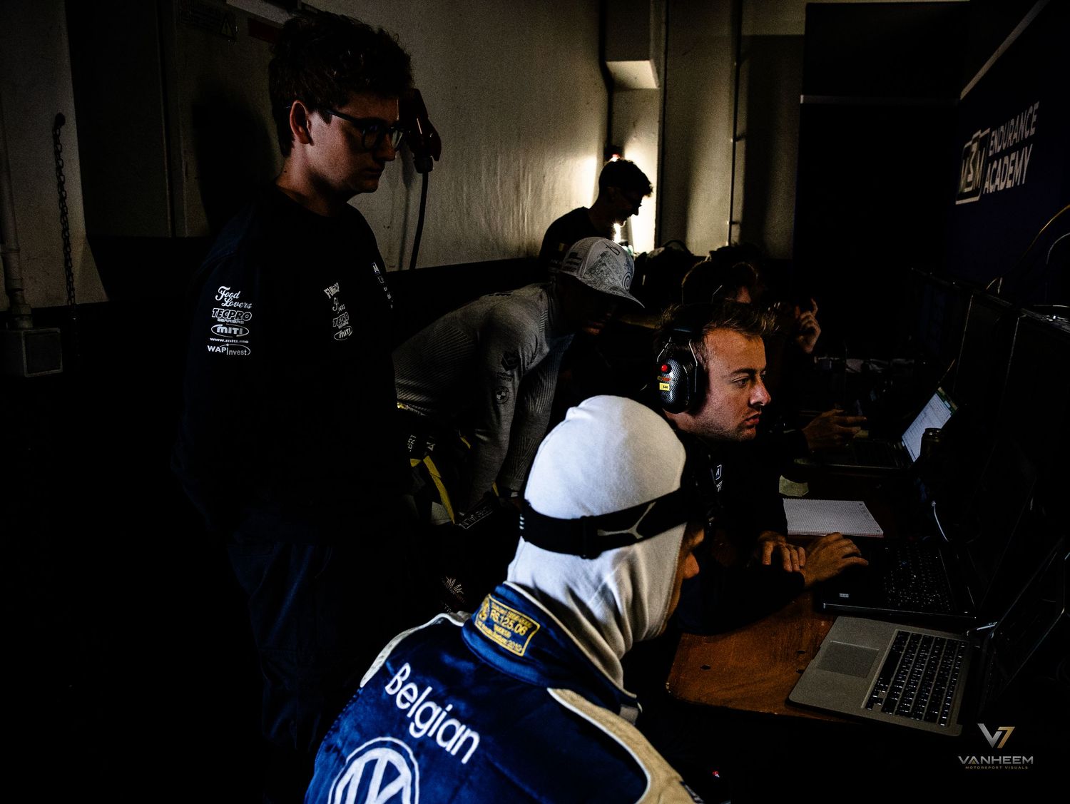 Racing team members analyze data on laptops in a dimly lit pit garage during a motorsport event.